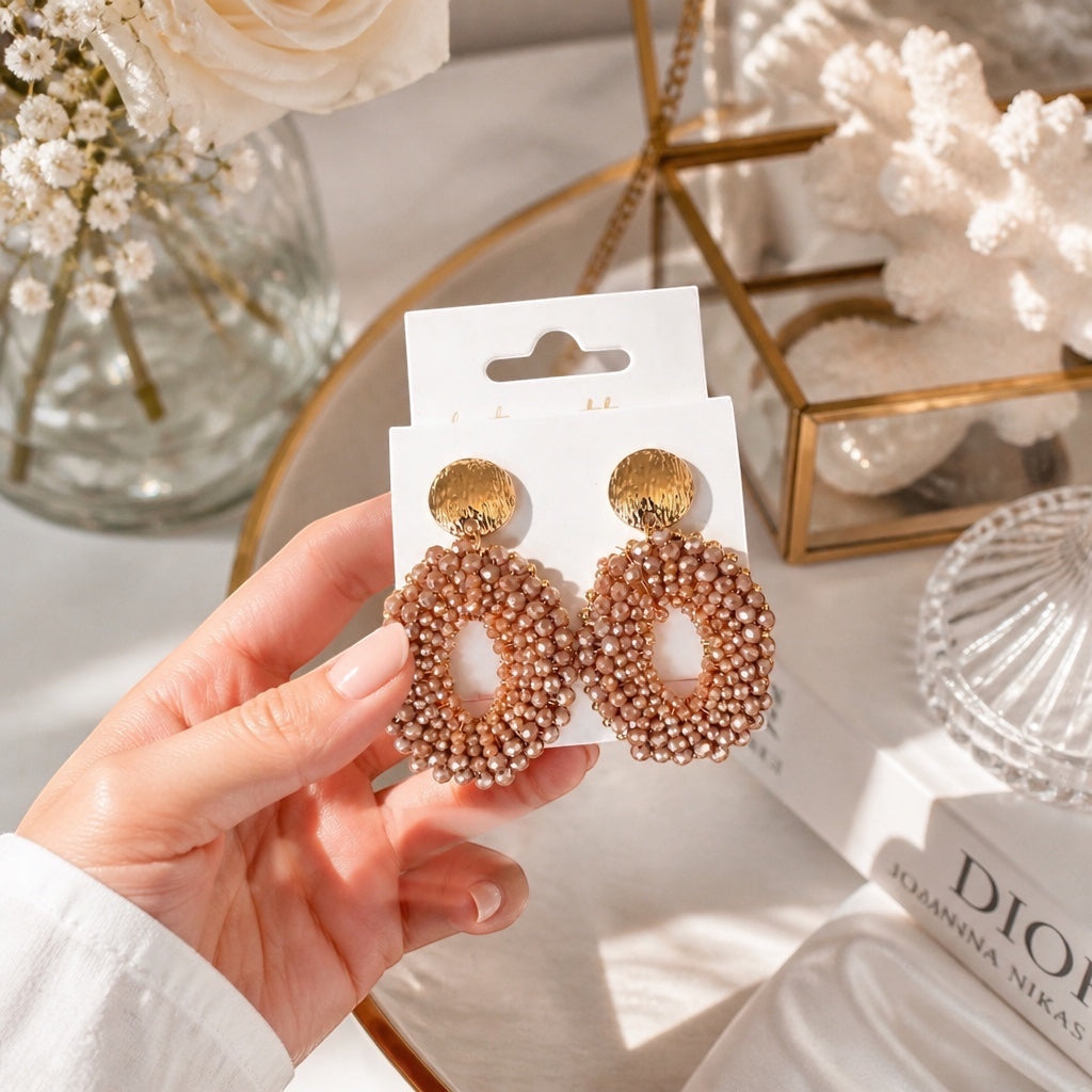 Home-Industry South Africa champagne beaded oval statement earrings with hammered gold studs held in a hand.